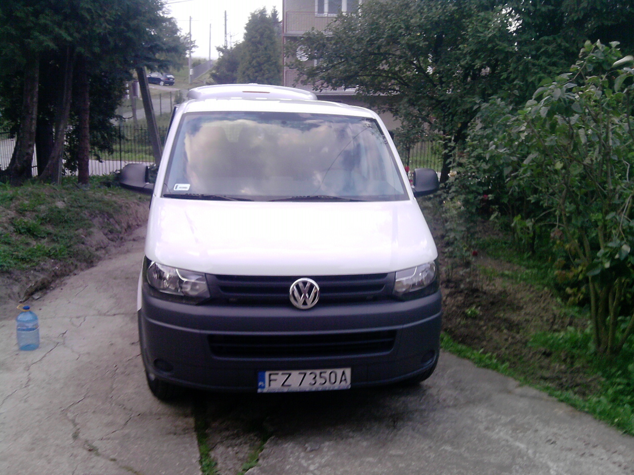 Biały Volkswagen Transporter zaparkowany na betonowej drodze w pobliżu zieleni, z widoczną butelką wody po lewej stronie.