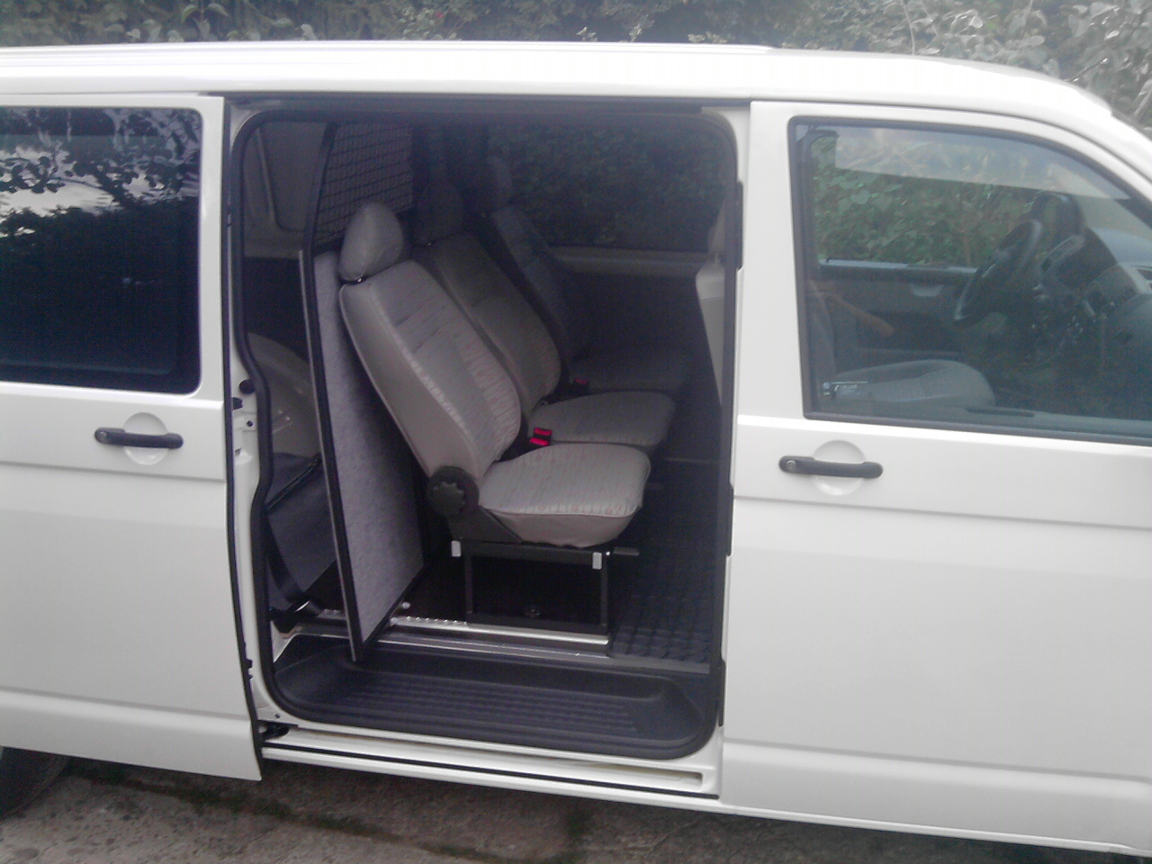 Wnętrze białego busa z otwartymi drzwiami przesuwnymi, widoczne szare fotele z zagłówkami i metalowa siatka zabezpieczająca przestrzeń pasażerską.