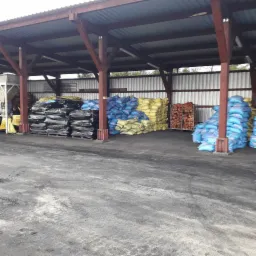 Zadaszony magazyn z zapasem opału: ułożone worki z materiałem opałowym w odcieniach czerni, błękitu i żółci oraz drewno kominkowe w metalowej klatce.