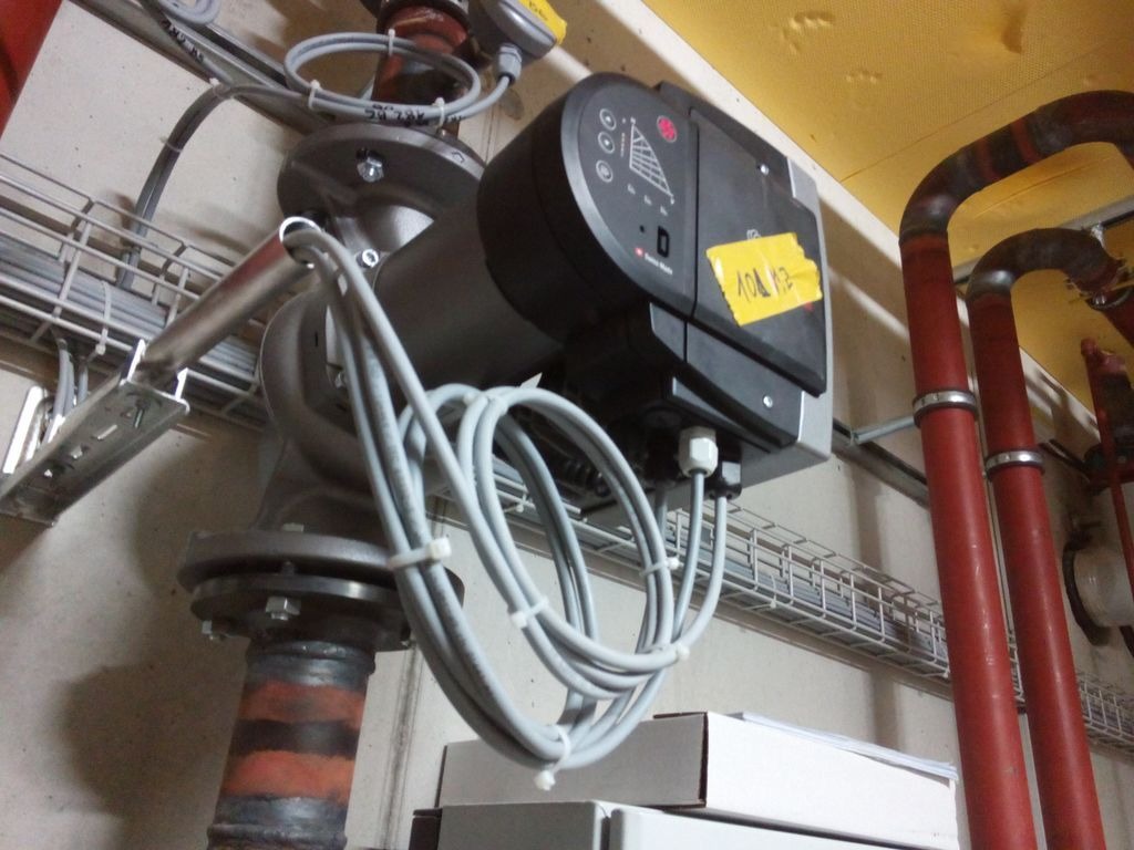 Szara pompa obiegowa zamontowana na rurociągu, z widocznymi przewodami elektrycznymi i żółtą naklejką z notatkami, na tle instalacji rurowej i metalowych korytek kablowych.