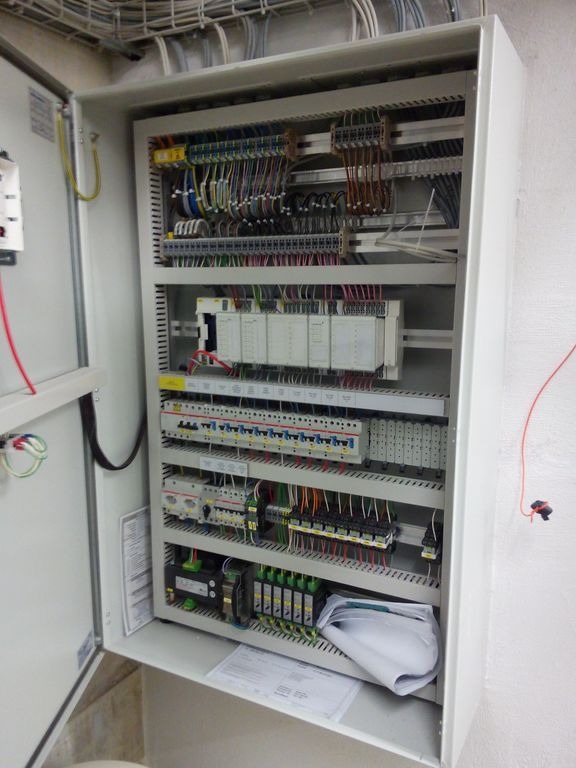 Otwarta szafa sterownicza z widocznymi przewodami, modułami sterującymi i zabezpieczeniami, umieszczona w pomieszczeniu technicznym z widocznymi kablami pod sufitem.