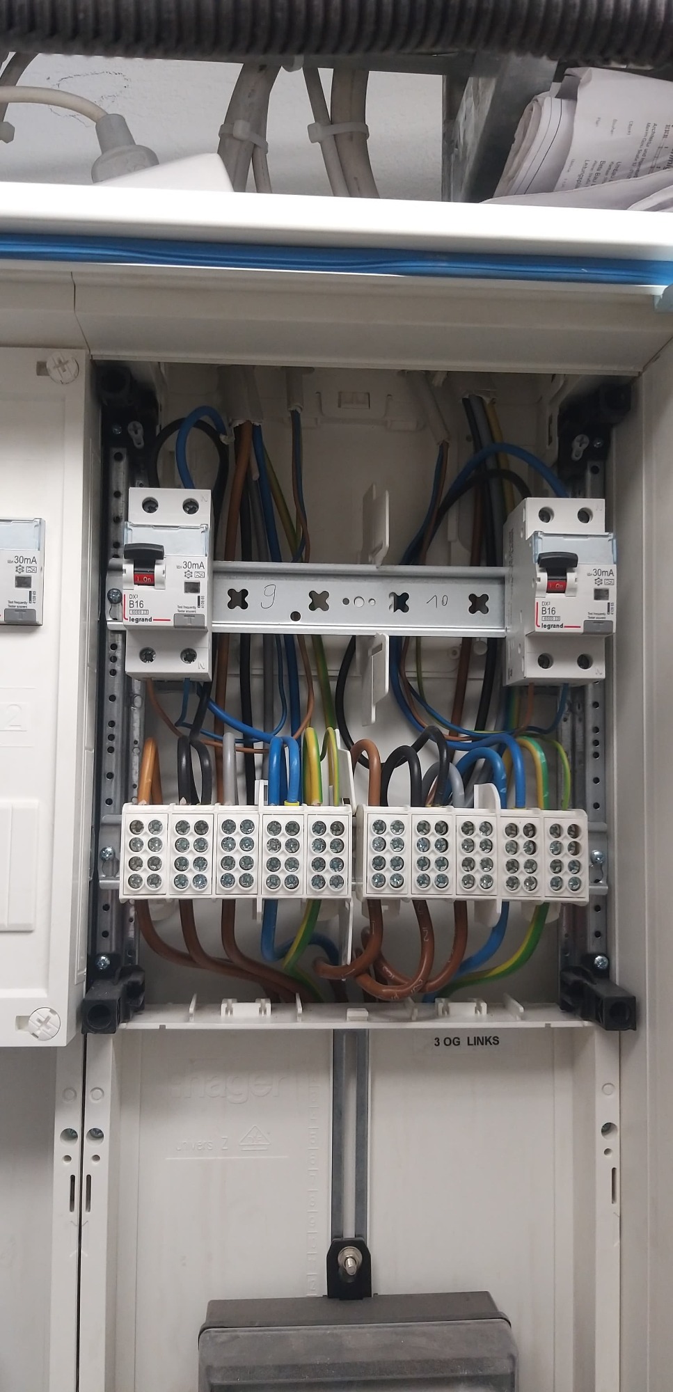 Otwarta skrzynka elektryczna z widocznymi bezpiecznikami Legrand, listwami zaciskowymi i uporządkowanymi przewodami w różnych kolorach, zamontowana w białej obudowie Hager.