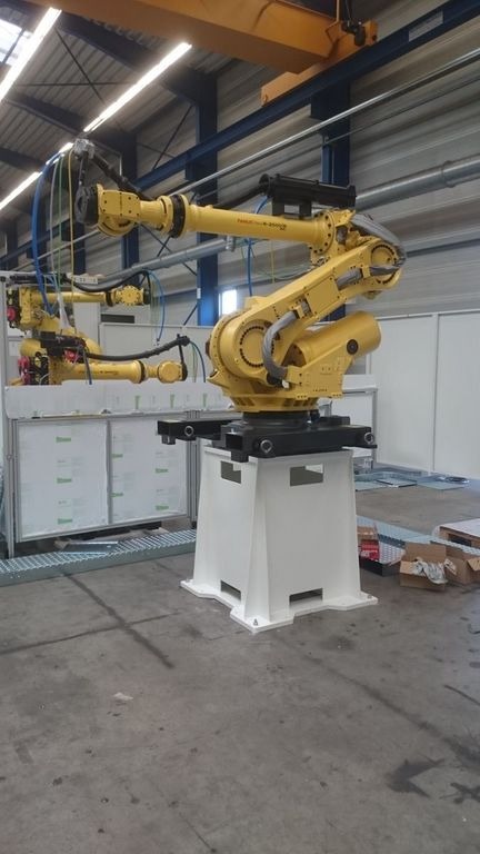 Żółty robot przemysłowy Fanuc R-2000iB na białej podstawie w hali produkcyjnej, z widocznymi przewodami pneumatycznymi i hydraulicznymi, oraz drugim robotem w tle.