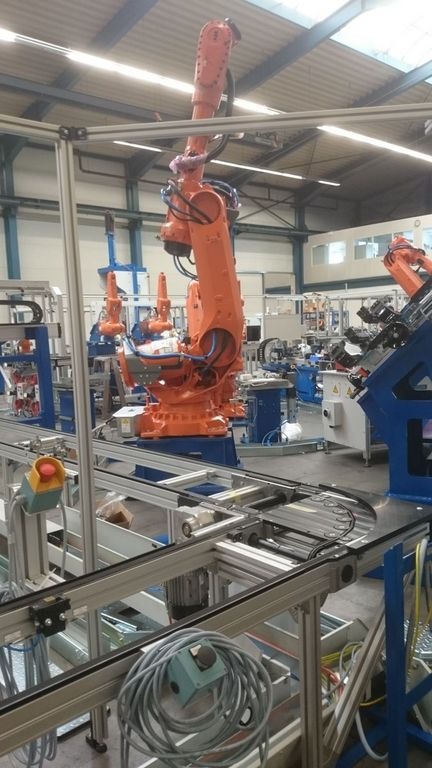 Pomarańczowy robot przemysłowy KUKA na niebieskiej podstawie, z widocznymi przewodami i elementami hydraulicznymi, w hali produkcyjnej z szynami transportowymi i innymi robotami w tle.