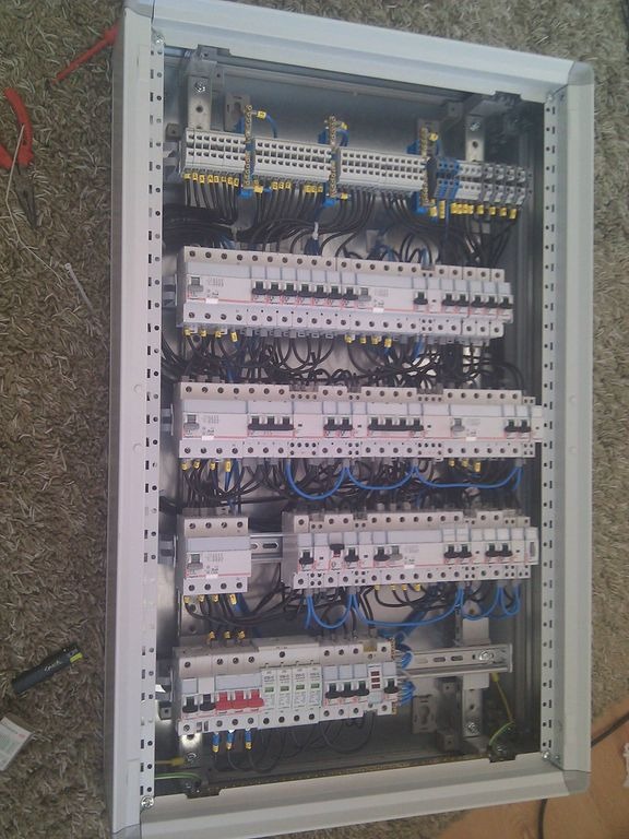 Rozdzielnica elektryczna w metalowej obudowie, widoczne zabezpieczenia różnicowoprądowe, bezpieczniki, listwy zaciskowe i okablowanie w kolorach czarnym i niebieskim, umieszczona na dywanie.