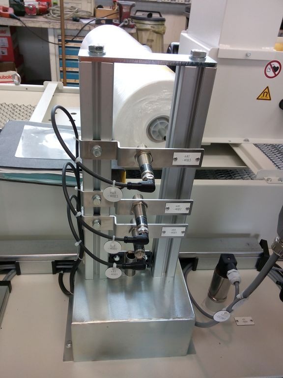 Szczegółowe zbliżenie na elementy automatyki przemysłowej: aluminiowa konstrukcja wsporcza z rolką folii, czujniki ciśnienia z przewodami i oznaczeniami identyfikacyjnymi, widoczne śruby mocujące...