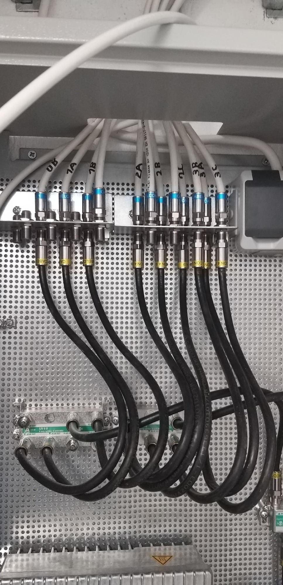 Rozdzielnica z licznymi kablami koncentrycznymi oznaczonymi numerami, podłączonymi do złączek na perforowanej metalowej płycie wewnątrz obudowy.