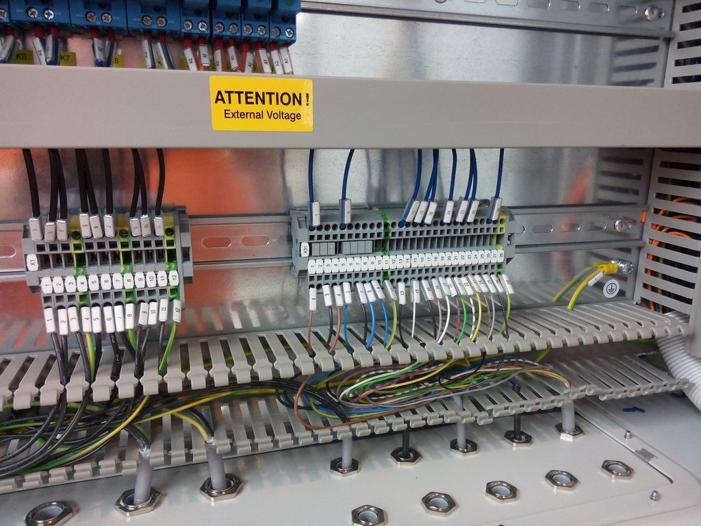 Wnętrze szafy sterowniczej z widocznymi listwami zaciskowymi, przewodami o różnych kolorach i naklejką ostrzegawczą 'Attention! External Voltage'.