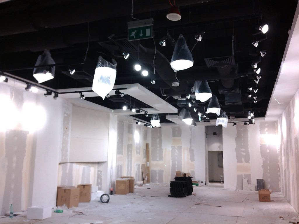 Wnętrze w trakcie prac wykończeniowych: surowa ściana z gipsem, podwieszany sufit z licznymi punktowymi lampami zabezpieczonymi folią, kartony na podłodze, widoczne kable i przewody.