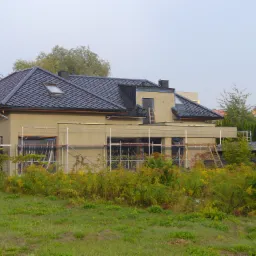 Nowoczesny dom z ciemną dachówką i oknami dachowymi w trakcie prac wykończeniowych, widoczne rusztowania i drabiny.