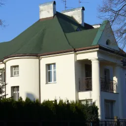 Dwupiętrowy dom z zielonym dachem z gontów bitumicznych i białymi ścianami, widoczne kominy z metalowymi nakładkami i antena