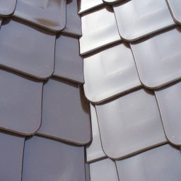 Agencja Budowlana WITAND - Błyszczące, brązowe dachówki bitumiczne ułożone w regularny wzór, widoczne z bliska pod kątem, z refleksami światła podkreślającymi ich gładką powierzchnię.