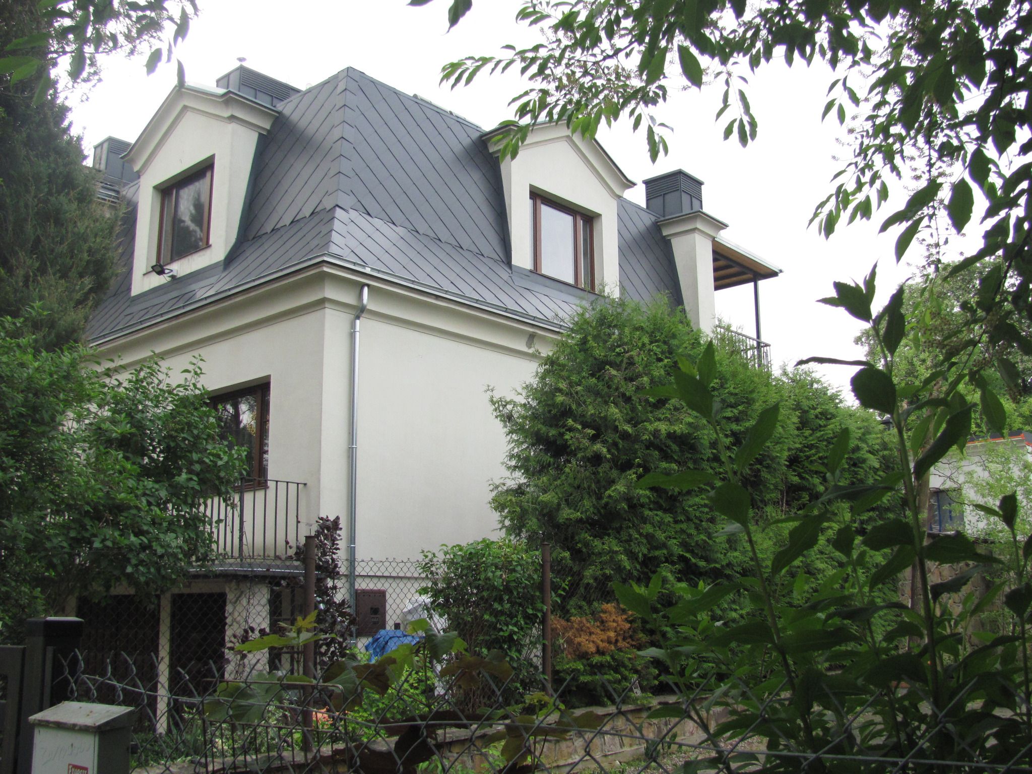 Elewacja domu z ciemnoszarym dachem z lukarnami, widoczna zza zieleni. Dachówka ułożona w regularny wzór. Jasna elewacja kontrastuje z ciemnym dachem.