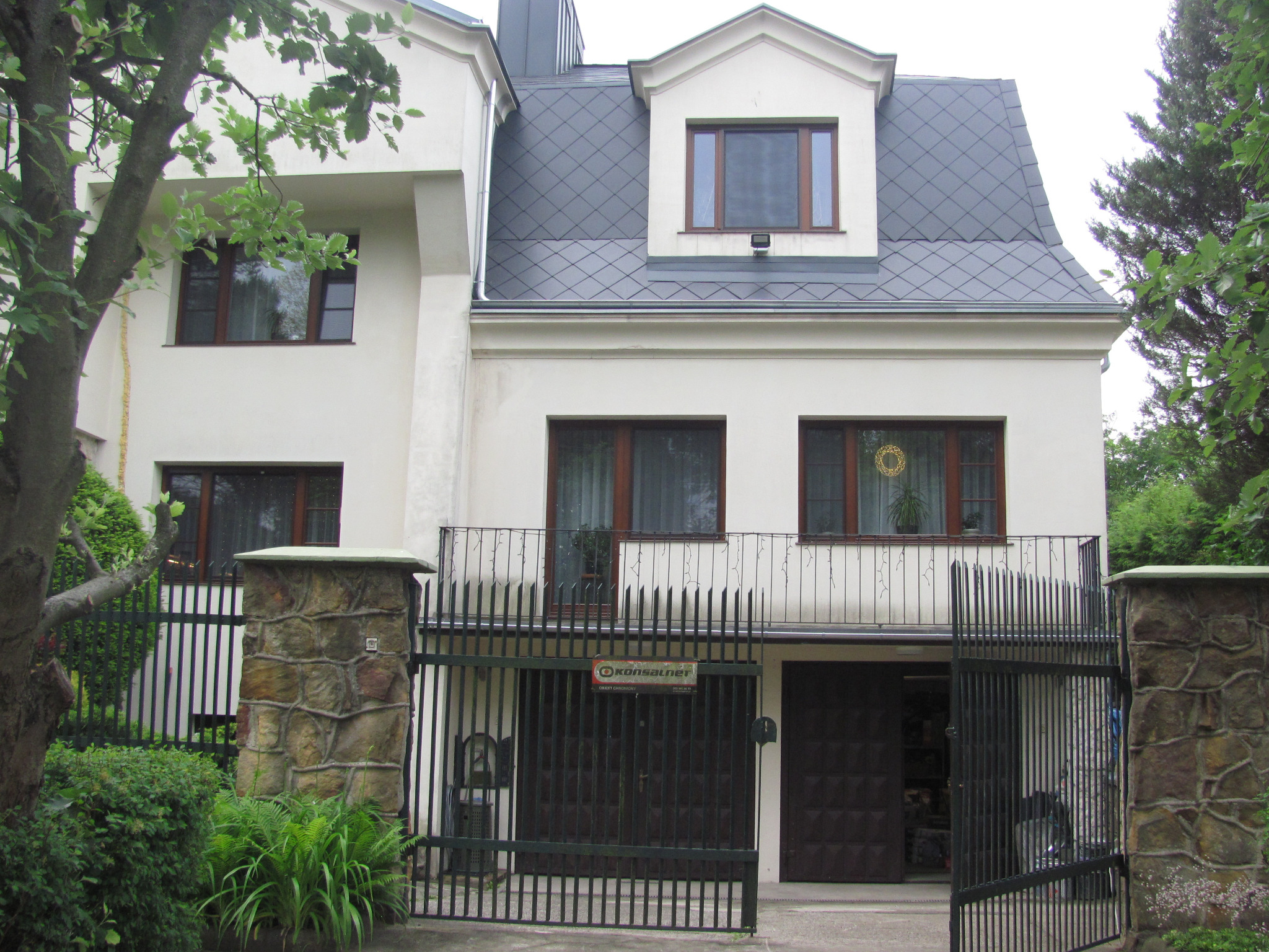 Dwupiętrowy dom z szarym dachem krytym romboidalną dachówką, widoczny zza czarnego, metalowego ogrodzenia z kamiennymi słupkami. Otwarta brama garażowa.