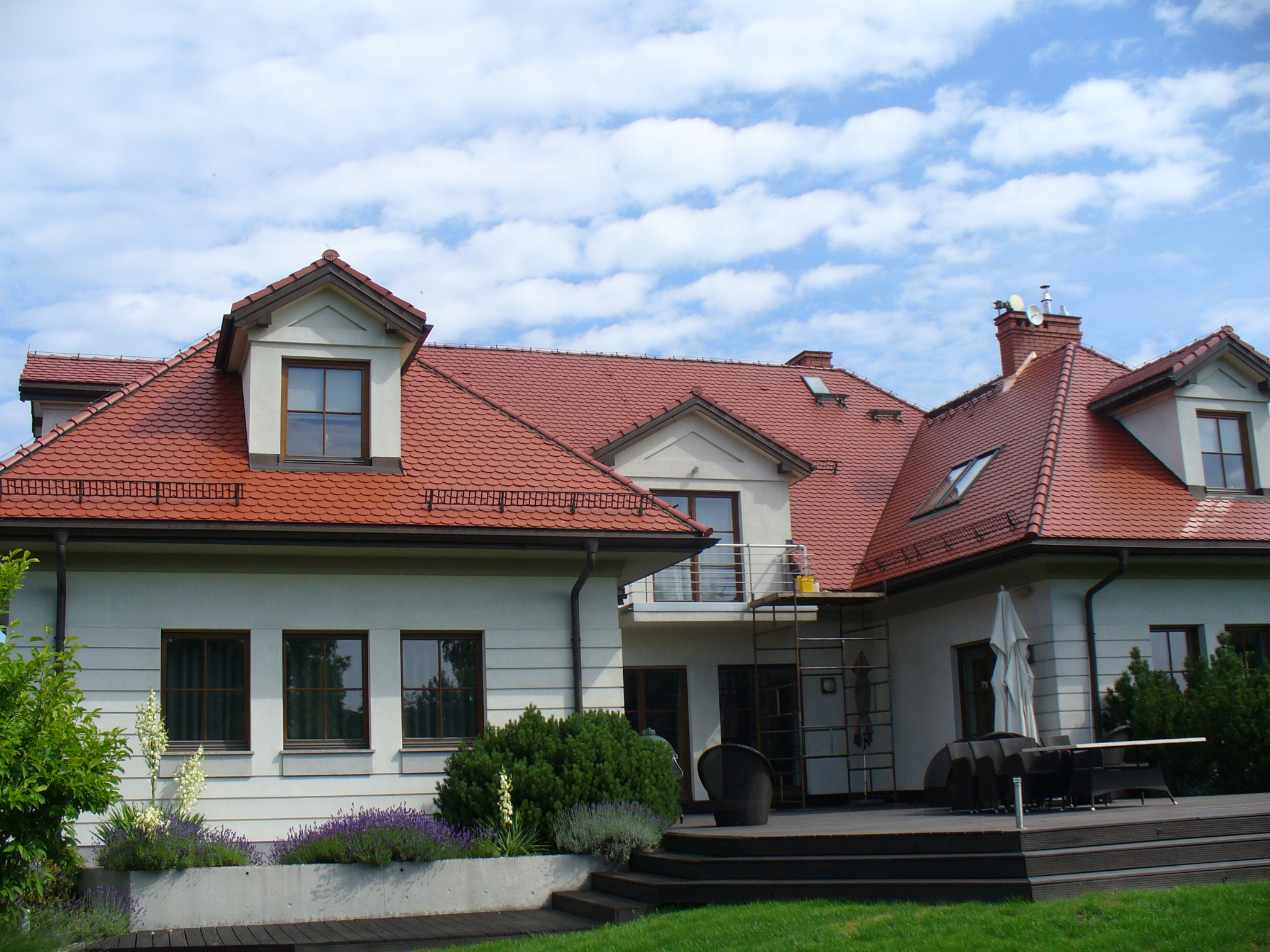 Rozległy dom jednorodzinny z czerwoną dachówką karpiówką, jasną elewacją i tarasem. Widoczne okna dachowe, komin i zadbany ogród z elementami architektury krajobrazu.