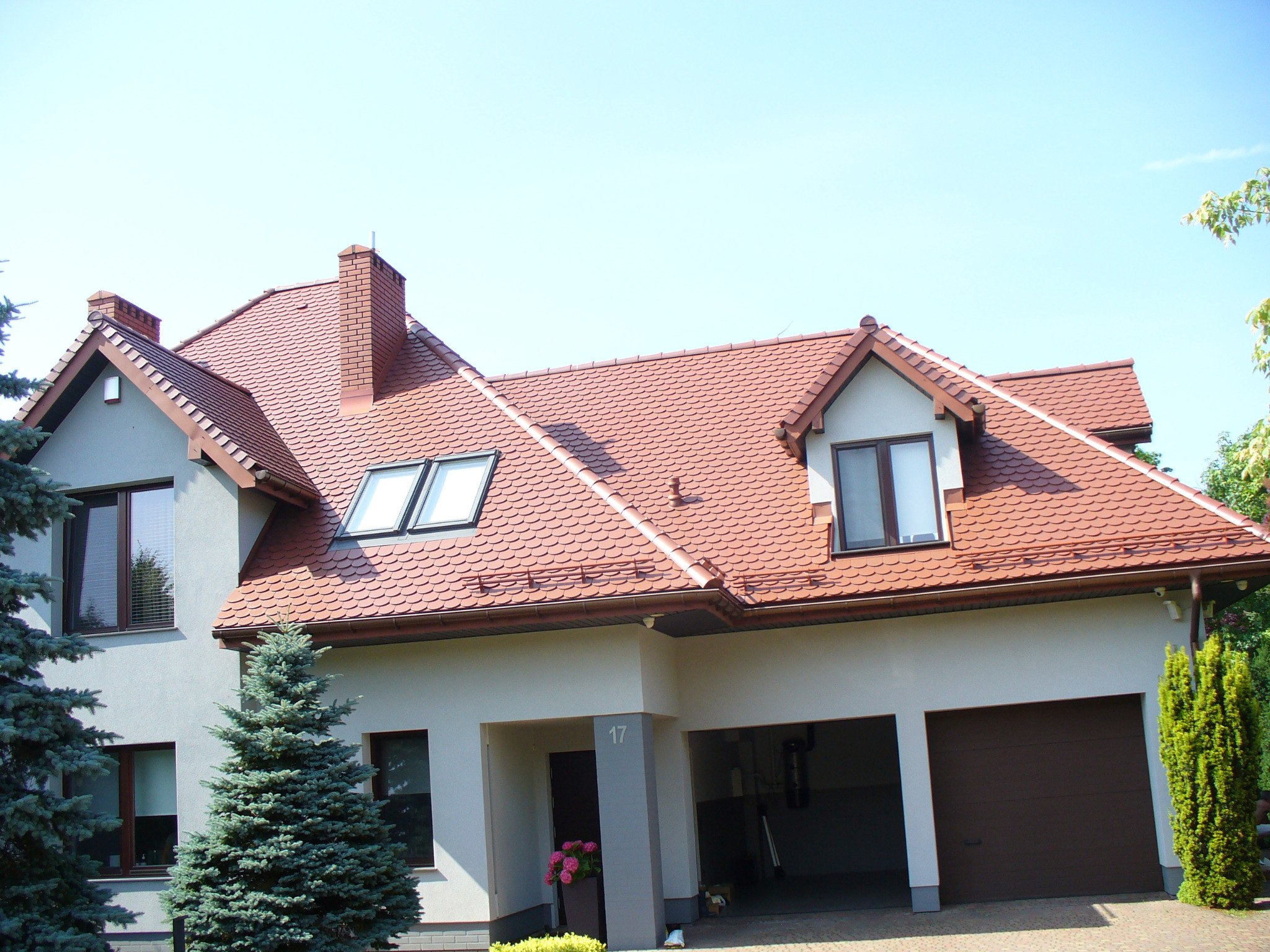 Dom jednorodzinny z dachem pokrytym czerwoną dachówką karpiówką. Widoczne okna dachowe, komin z cegły i zadbany ogród z iglakami.