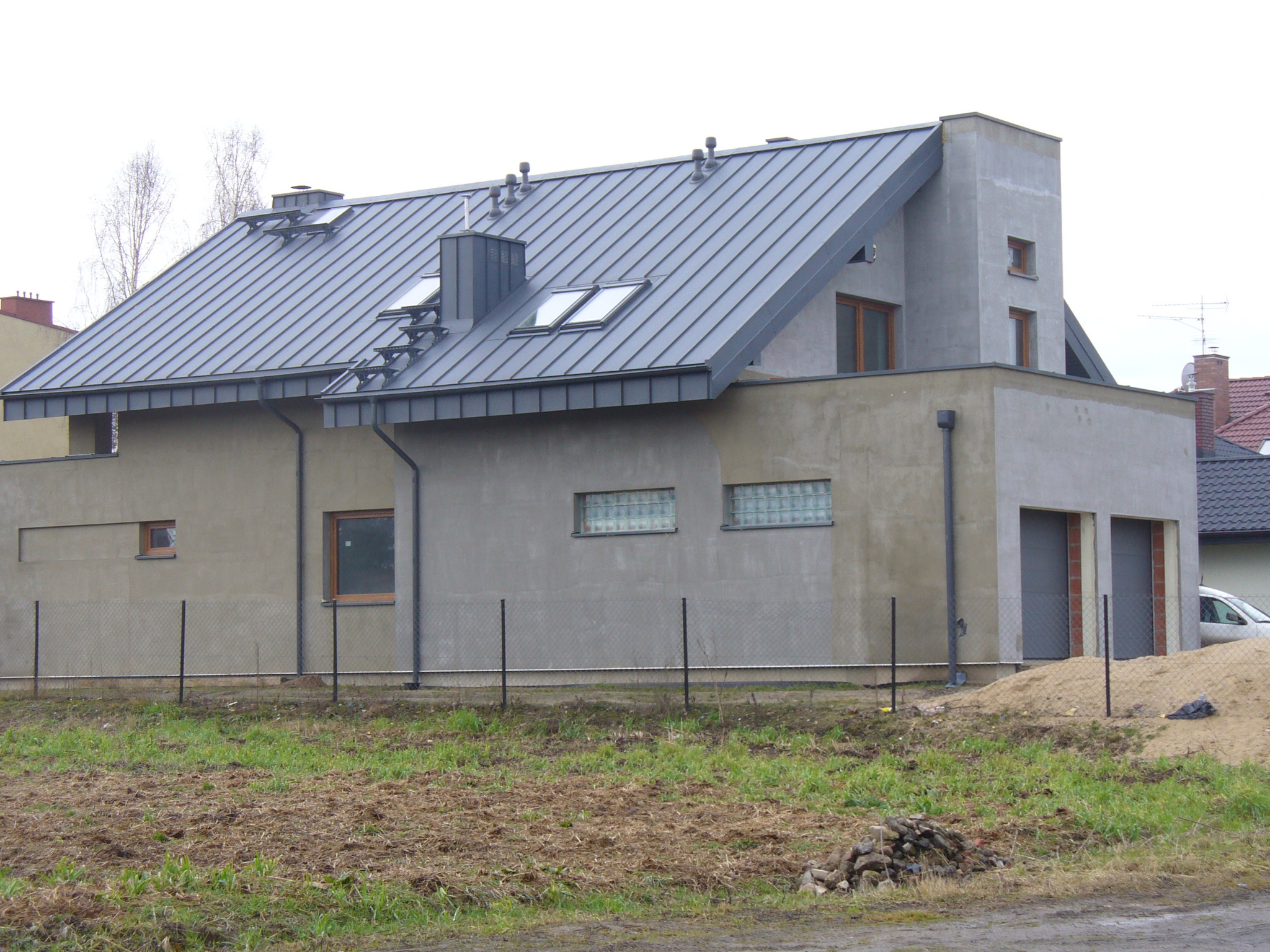 Nowoczesny dom jednorodzinny z szarym dachem z blachy na rąbek stojący, okna dachowe, elewacja w odcieniach szarości, widok z zewnątrz, ogrodzenie z siatki.