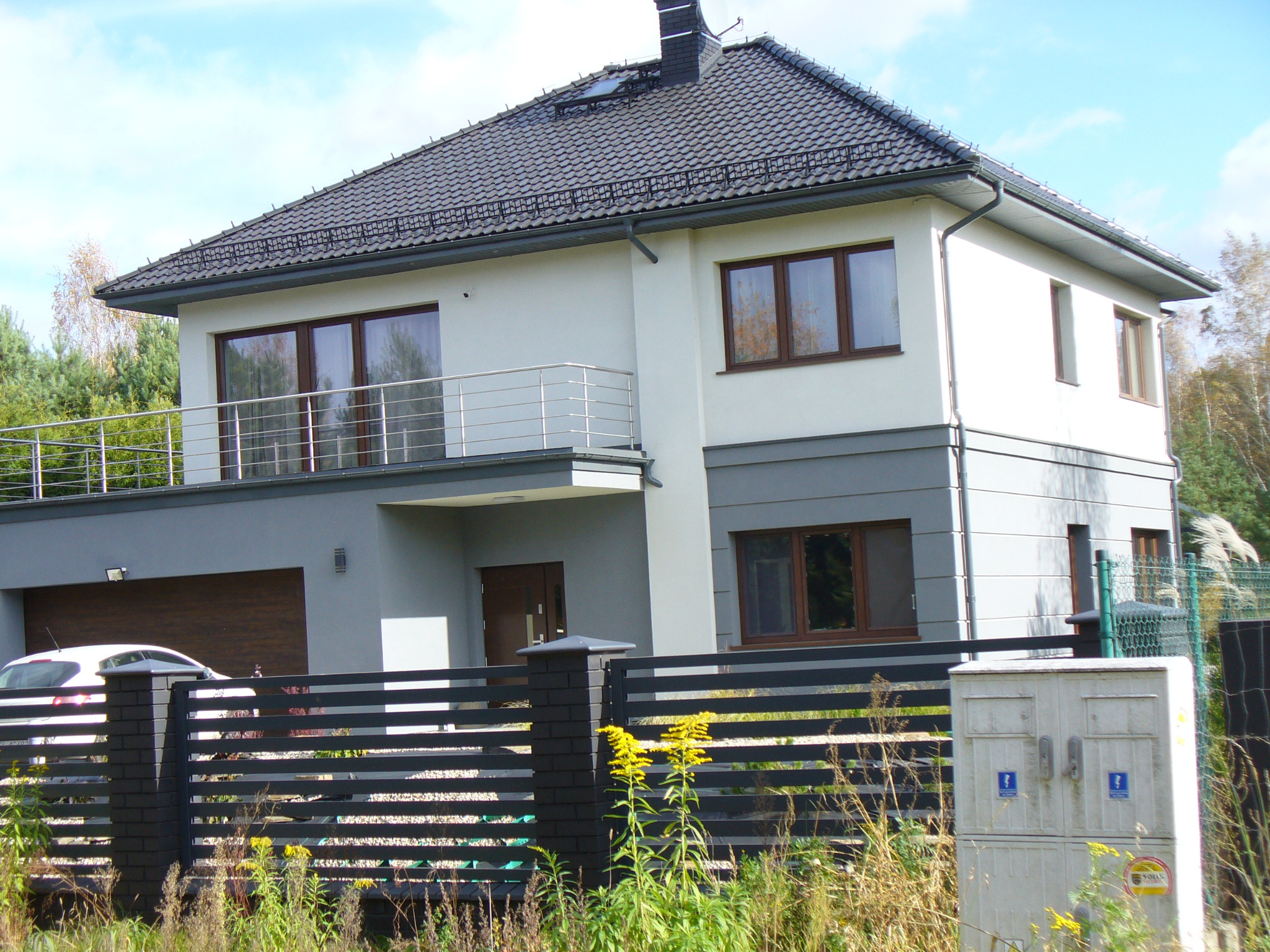Dwukondygnacyjny dom z ciemnoszarym dachem i balkonem, otoczony czarnym ogrodzeniem. Widoczne okna z brązowymi ramami i garaż z bramą segmentową.