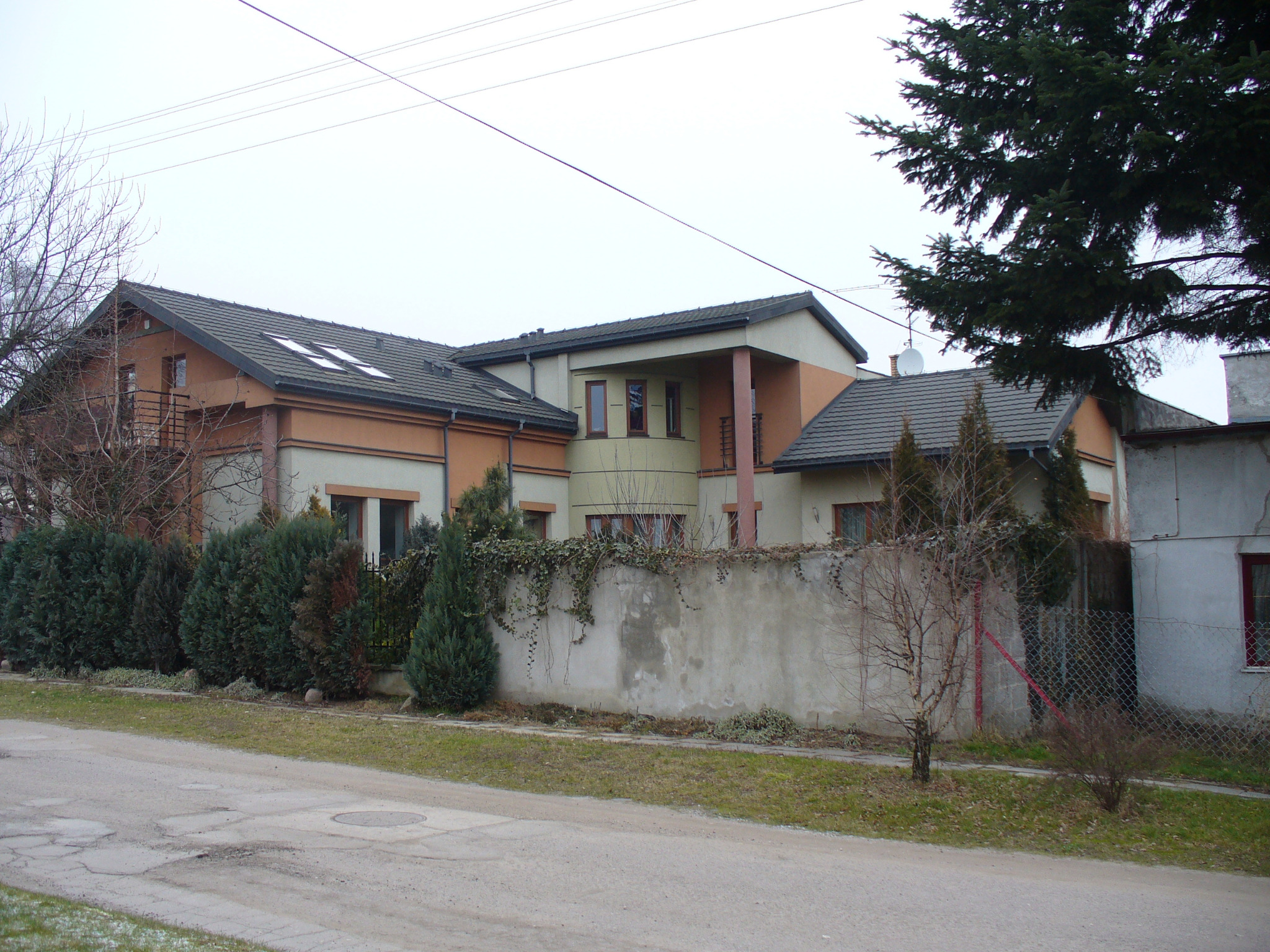 Dwukondygnacyjny dom z szarym dachem z dachówki, widoczny z ulicy. Na dachu okna połaciowe. Część elewacji w kolorze beżowym, część w pomarańczowym.