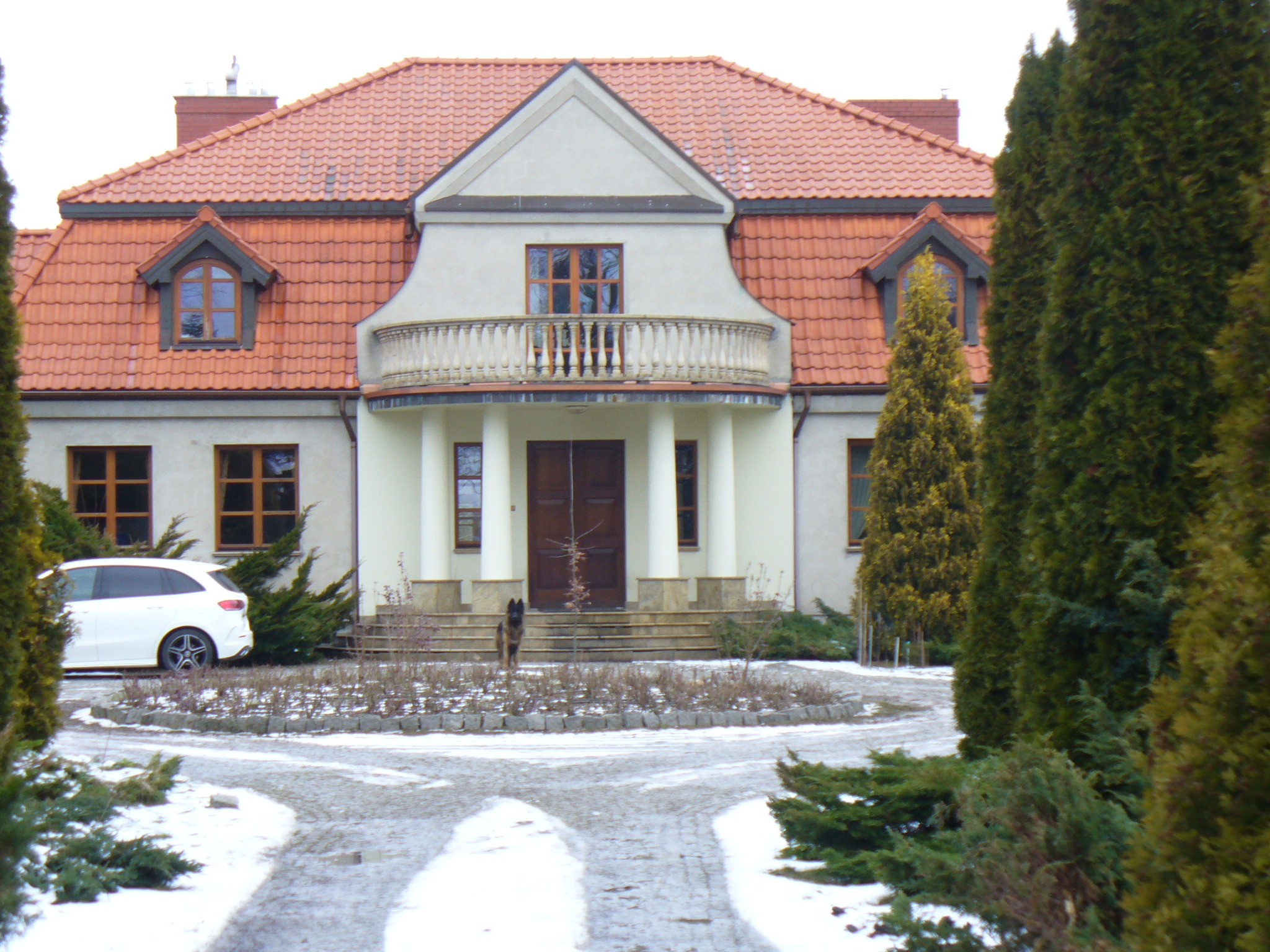 Dwupiętrowy dom z czerwonym dachem, balkonem i kolumnami. Na podjeździe biały samochód i pies. Zima, śnieg na trawniku i ścieżce.