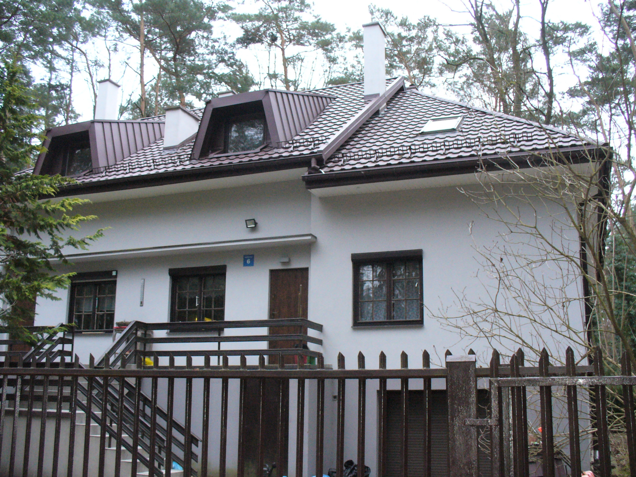 Dwupiętrowy dom z brązowym dachem z dachówką, widoczny zza drewnianego ogrodzenia. Dach ma okna dachowe i kominy. W tle drzewa.