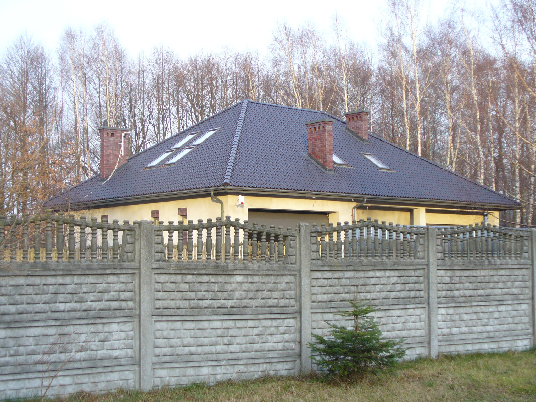 Dom jednorodzinny z ciemnym dachem z dachówką, ceglanymi kominami i oknami dachowymi, otoczony betonowym ogrodzeniem z dekoracyjnymi elementami.