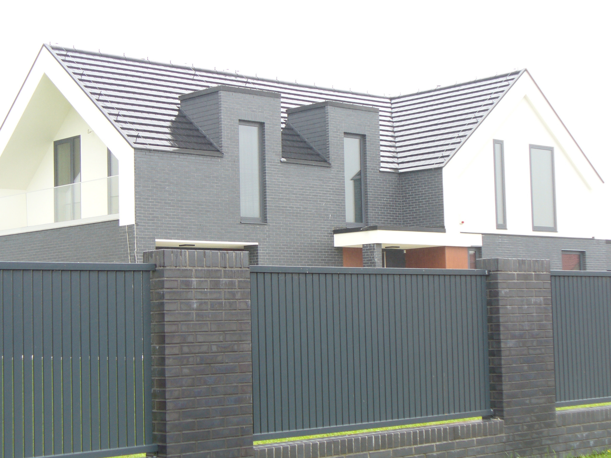 Nowoczesny dom jednorodzinny z szarej cegły, z ciemną dachówką i metalowym ogrodzeniem panelowym. Widok z poziomu gruntu, podkreślający geometrię budynku.