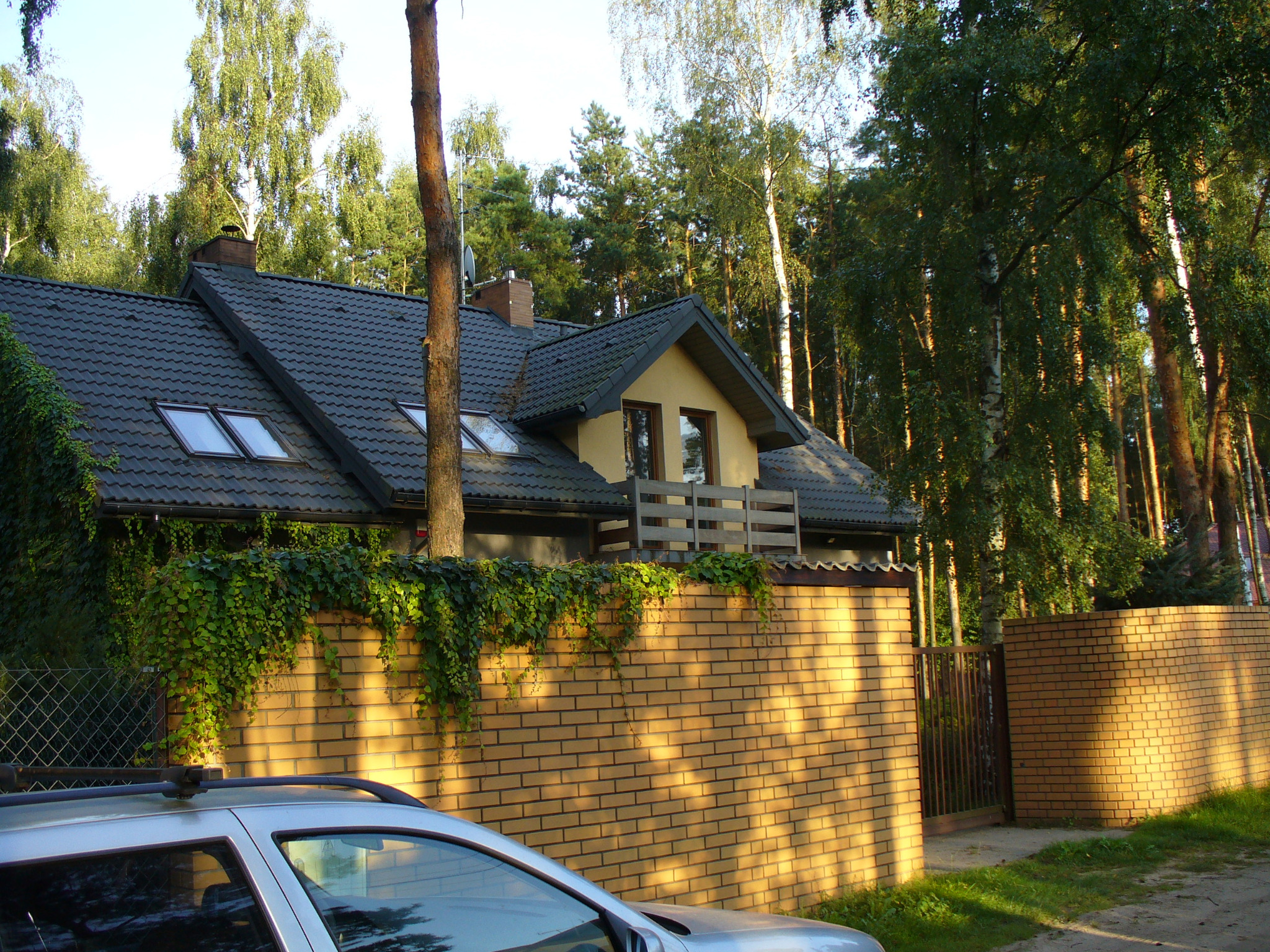 Dom z ciemną dachówką i oknami dachowymi, otoczony zielenią i ceglanym murem. Fragment samochodu w kadrze. Ujęcie w słoneczny dzień.