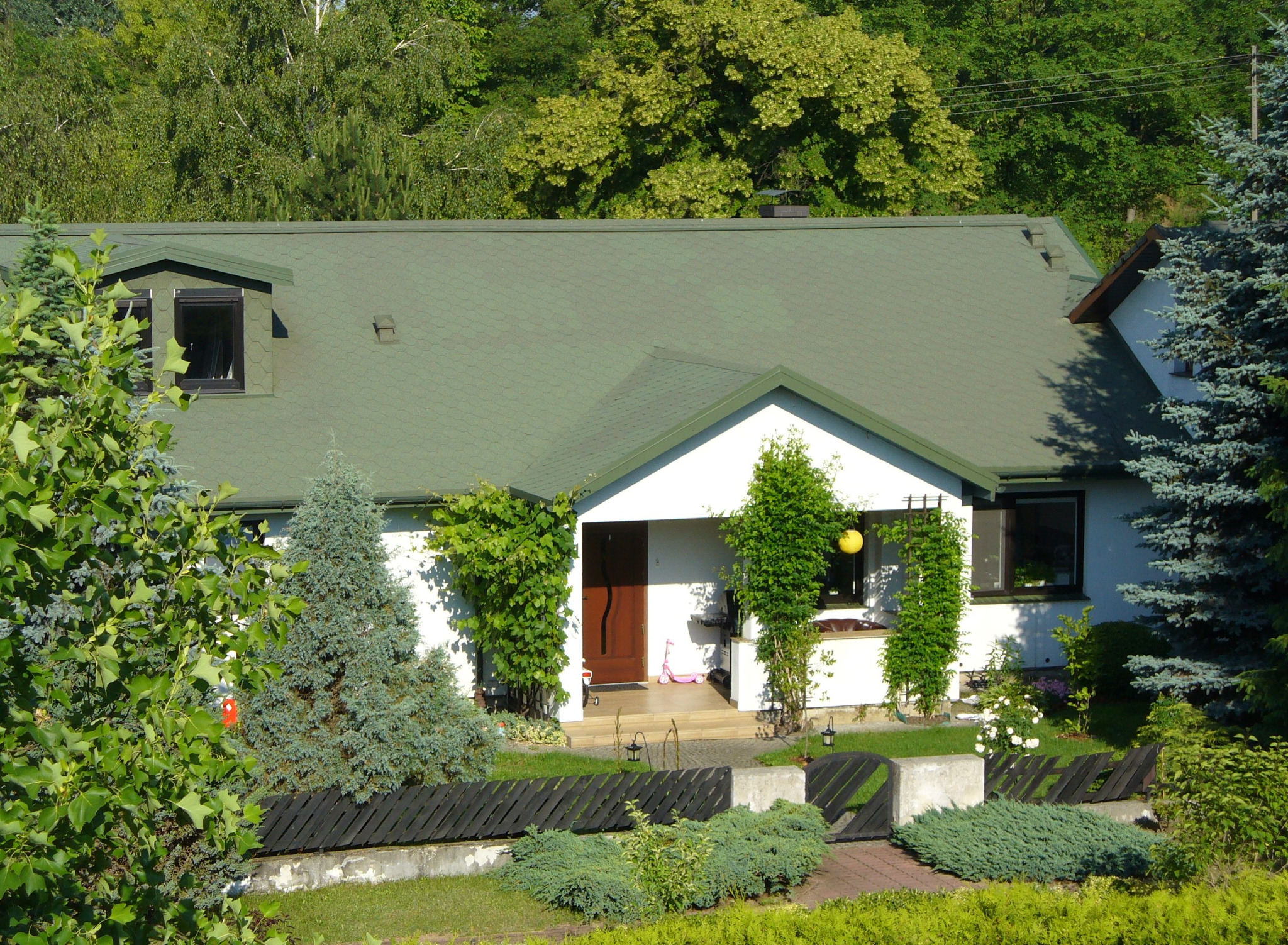 Dom jednorodzinny z zielonym dachem z dachówki bitumicznej, widok z przodu, otoczony zielenią. Na dachu widoczne okno dachowe. Uwagę przykuwa zadbany ogród.