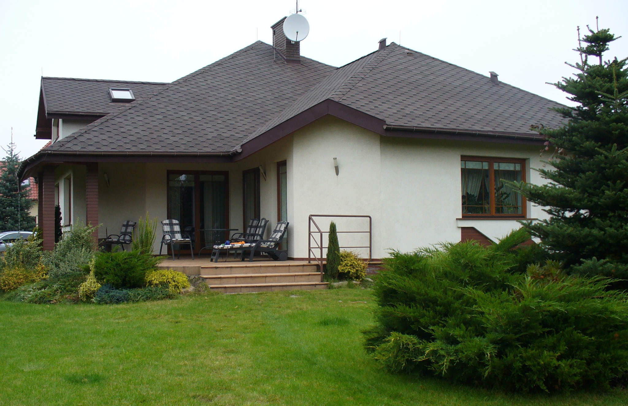 Dom jednorodzinny z brązowym dachem z gontu bitumicznego i anteną satelitarną. Widok z ogrodu, zieleń i elementy architektury ogrodowej.