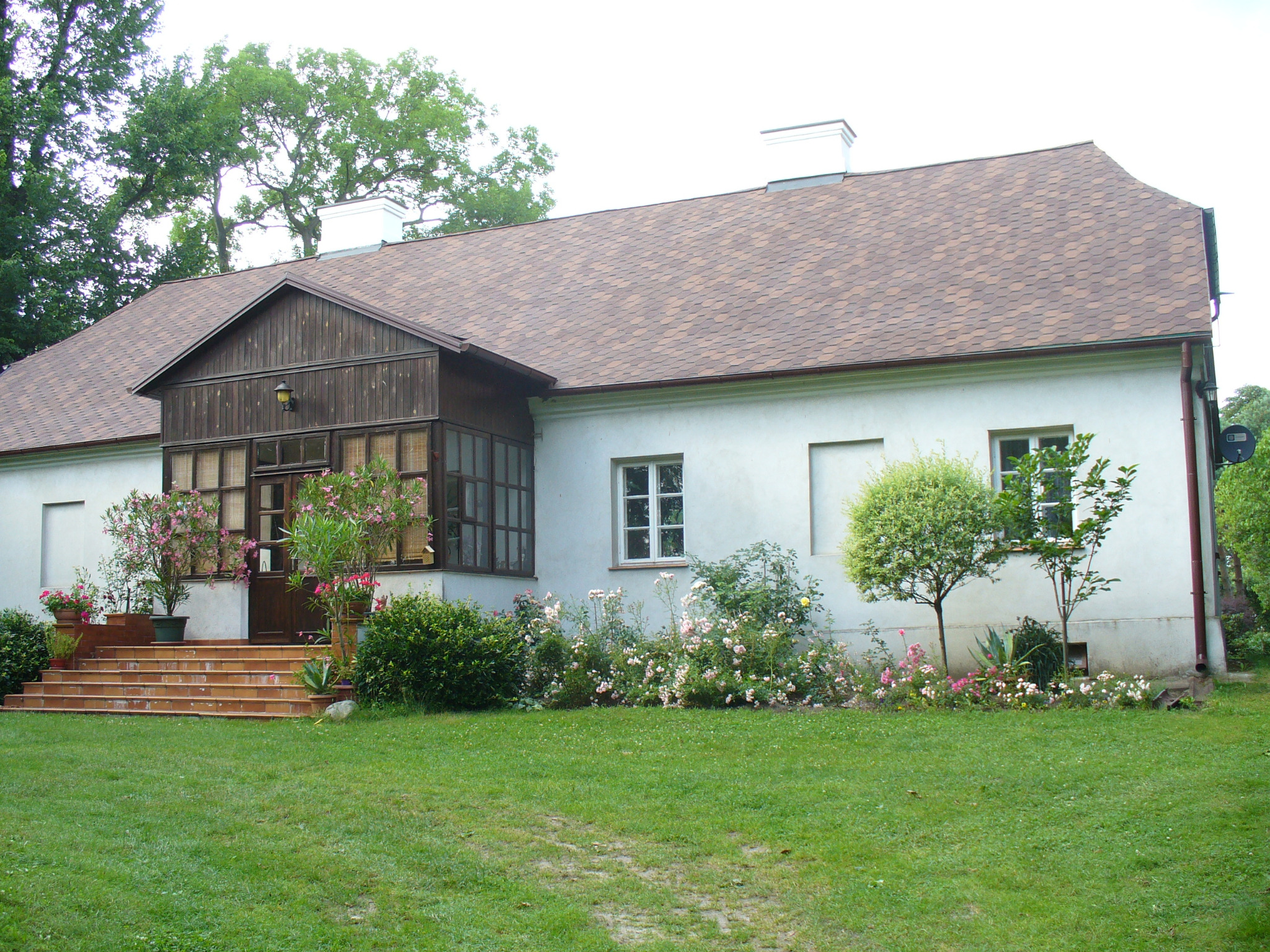 Rozległy parterowy dom z brązowym dachem z gontów bitumicznych, białą elewacją i werandą. Uroku dodają kwiaty i zadbany trawnik.