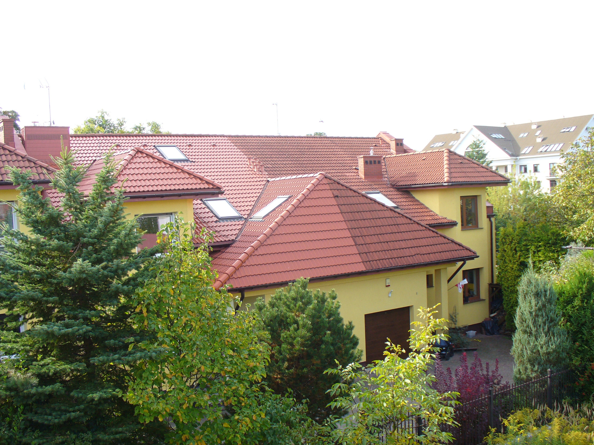 Żółty dom jednorodzinny z czerwoną dachówką, widok z góry, otoczony zielenią. Na dachu okna połaciowe i komin. W tle budynek wielorodzinny.