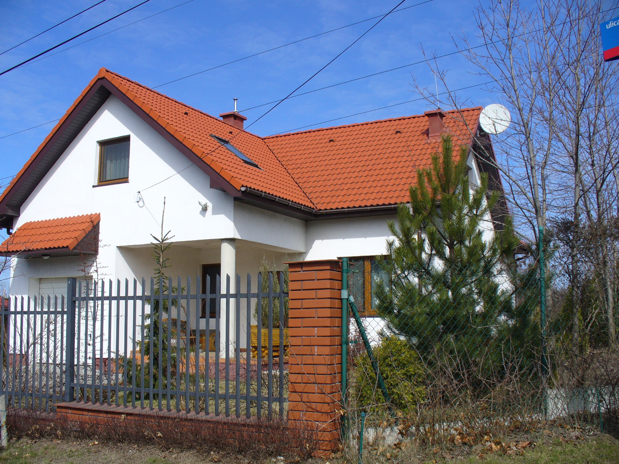Dom jednorodzinny z pomarańczową dachówką, widoczny zza metalowego ogrodzenia i zieleni. Na dachu antena satelitarna i okno dachowe.