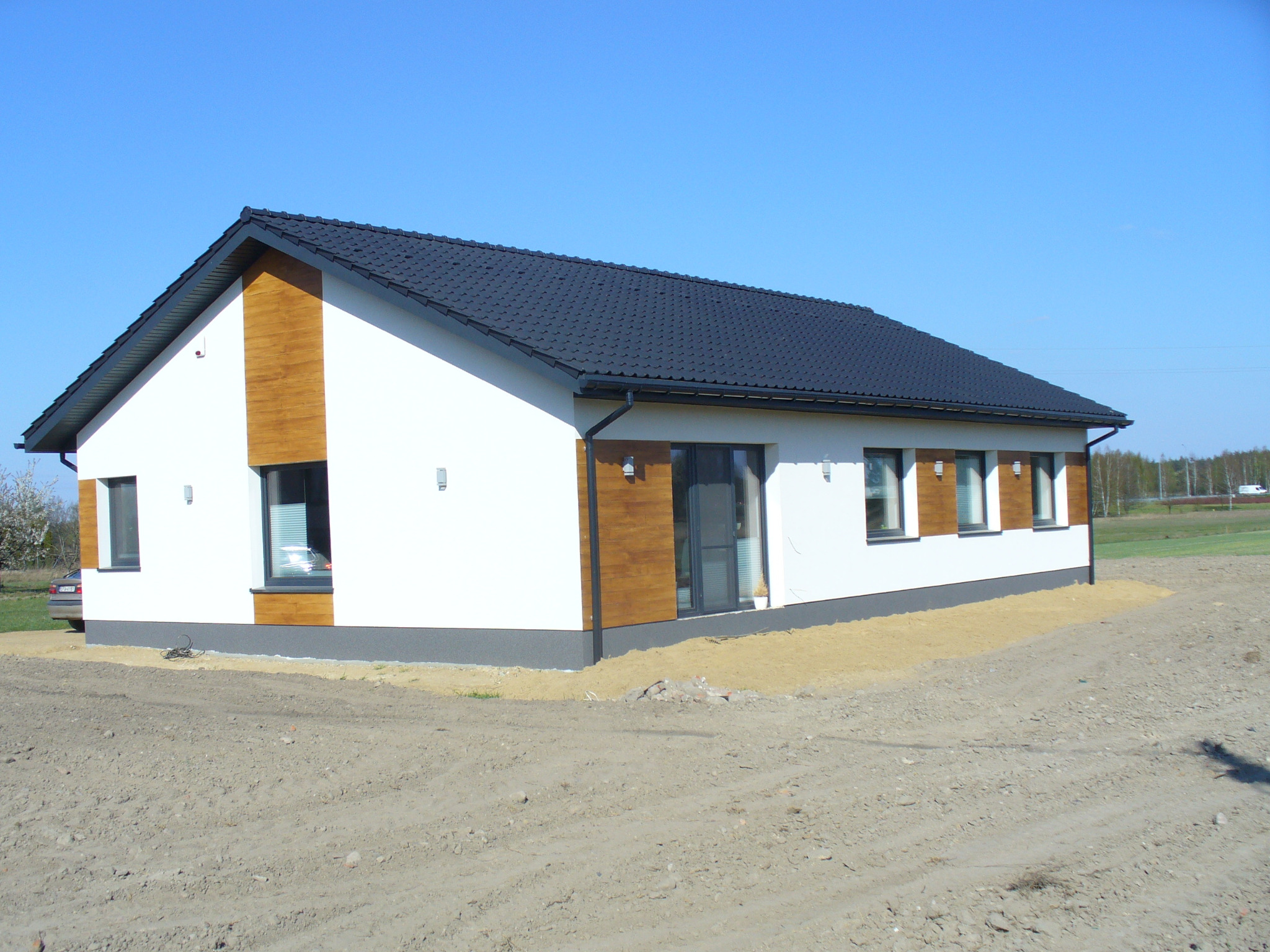 Nowoczesny dom jednorodzinny z czarnym dachem w Łodzi. Elewacja biała z drewnianymi akcentami. Widoczne okna i drzwi balkonowe. Teren wokół domu przygotowany pod trawnik.