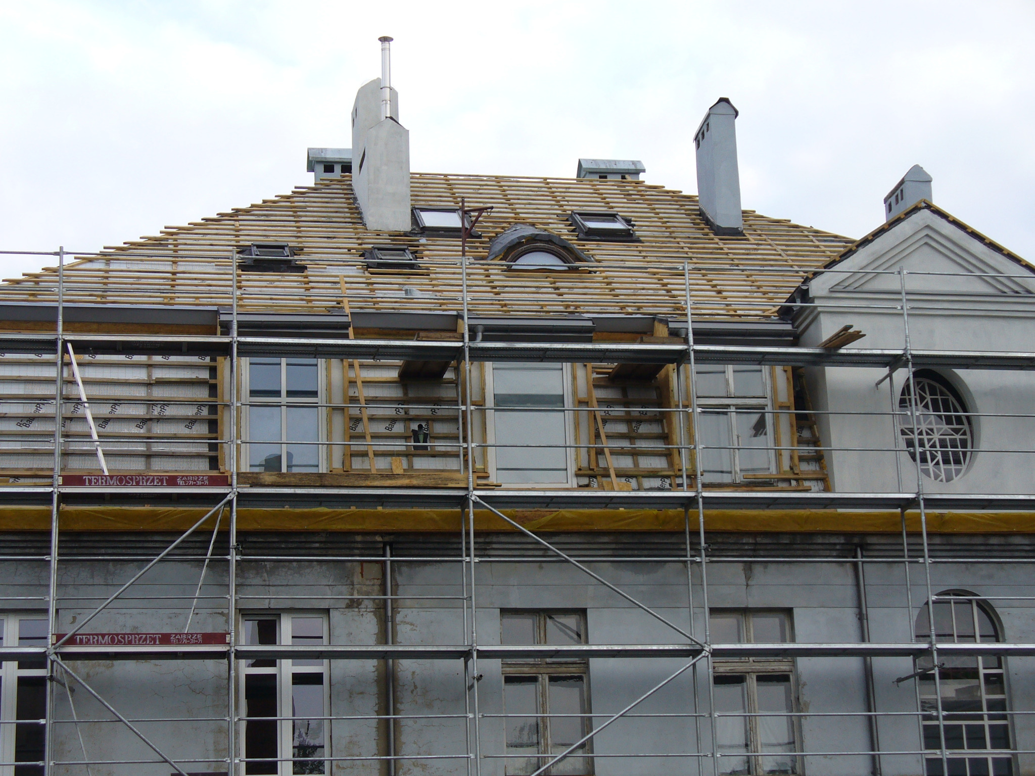 Budynek w rusztowaniach z odsłoniętą konstrukcją dachu, widoczne okna dachowe i kominy. Etap prac przed położeniem pokrycia dachowego.