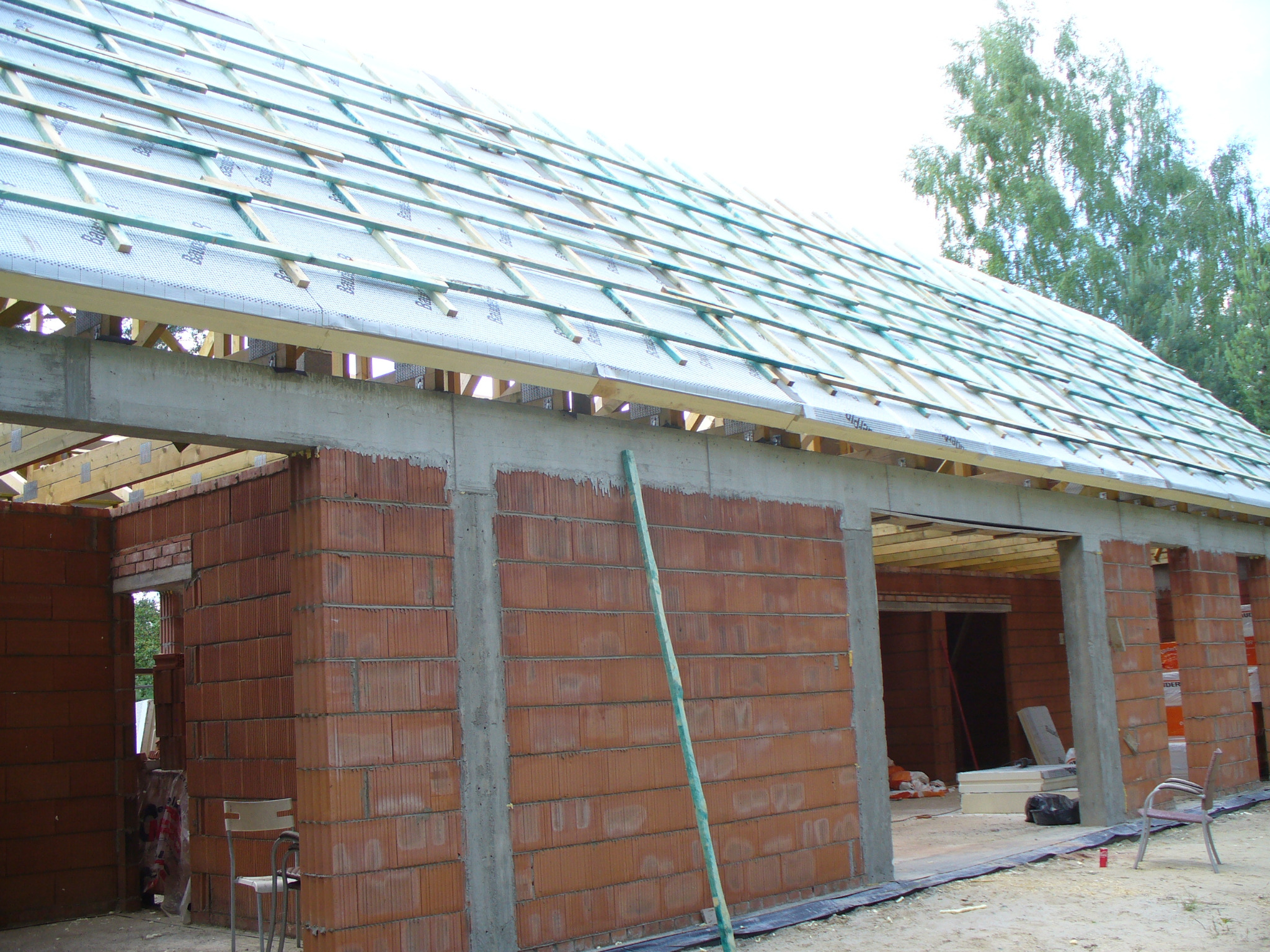 Budowa domu z czerwonej cegły: widok na konstrukcję dachu z membraną i łatami, betonowe nadproża, częściowo wzniesione ściany. W tle zieleń drzew.