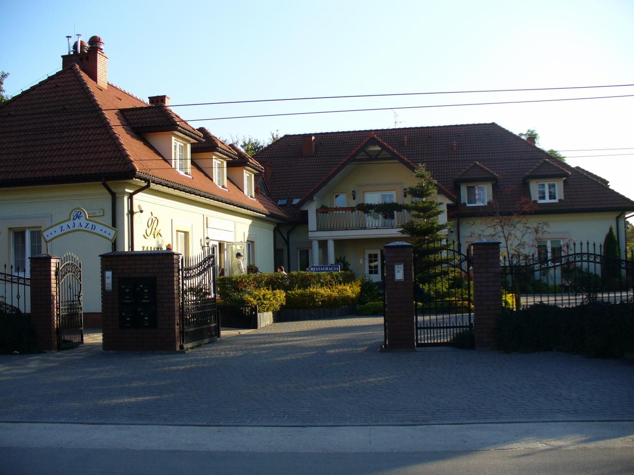 Dwukondygnacyjny budynek z czerwonym dachem, widoczny zza ozdobnego, kutego ogrodzenia. Dachówka ceramiczna, kominy z cegły. Budynek w stylu dworkowym.