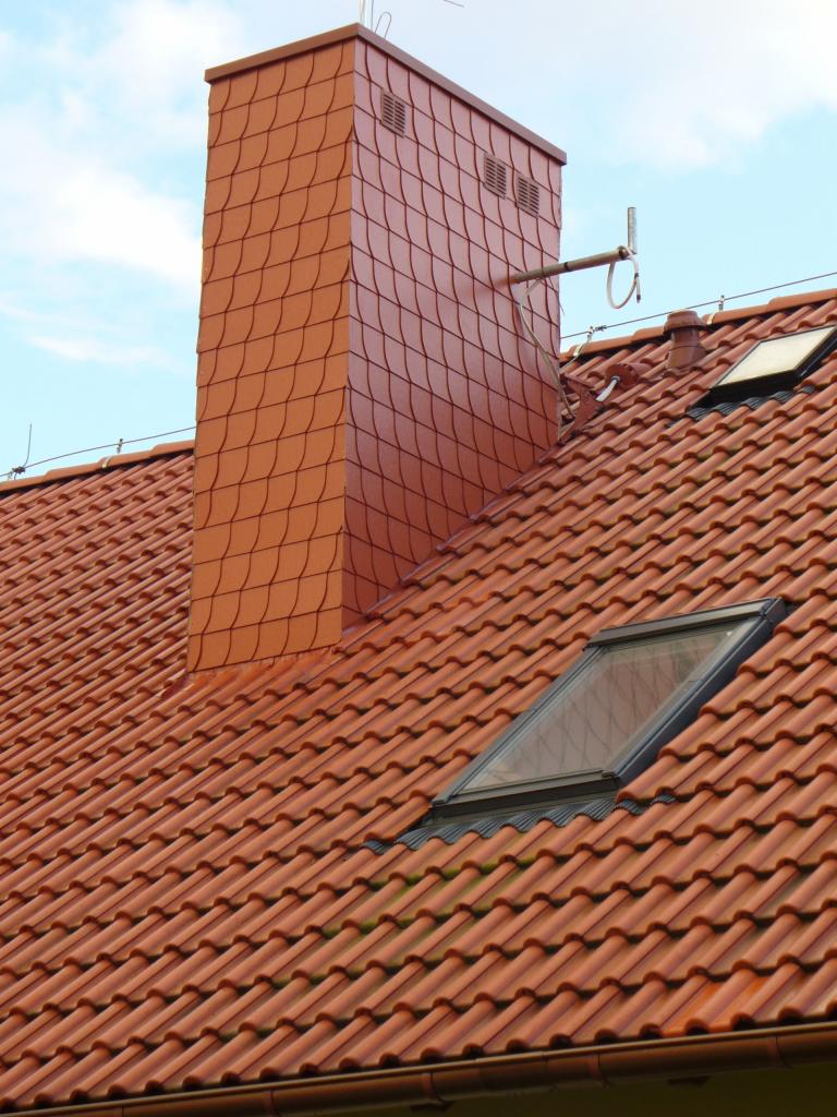 Czerwony dach z dachówką ceramiczną, komin obłożony łuską ceramiczną i dwa okna dachowe.
