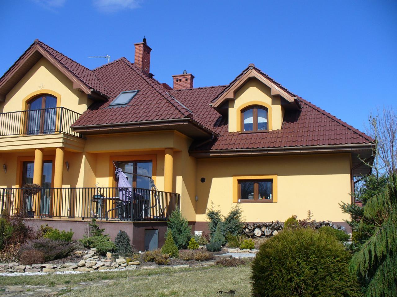 Żółty dom z brązowym dachem krytym dachówką, widok z ogrodu w słoneczny dzień. Balkony z czarną balustradą. Na dachu okno i komin.