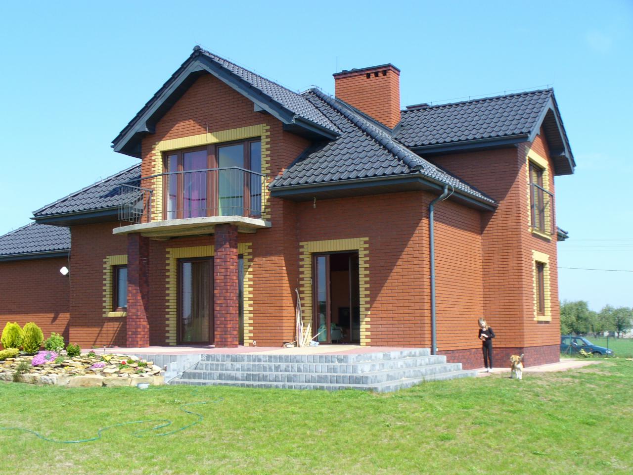 Dwupiętrowy dom z czerwonej cegły z ciemnoszarym dachem i balkonem. Widoczna kobieta z psem na trawniku. Elementy dekoracyjne w kolorze żółtym.