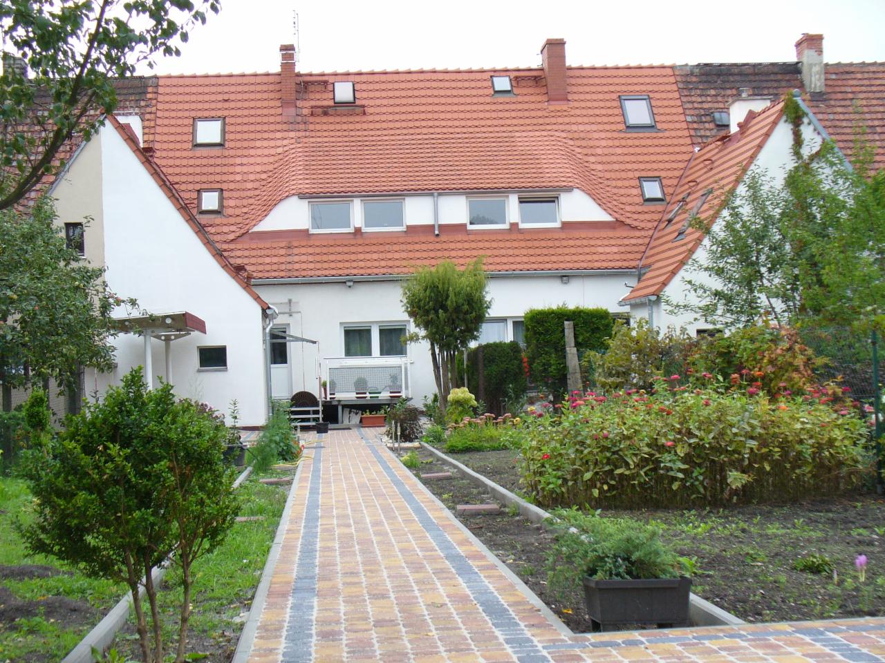 Widok na dom z czerwonym dachem z licznymi oknami dachowymi, ogród z brukowaną ścieżką i roślinnością. Ujęcie z poziomu ogrodu, w dzień.