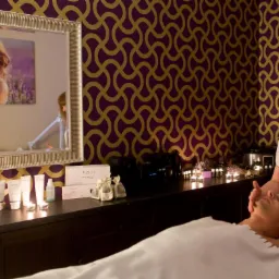 Mężczyzna relaksuje się podczas masażu twarzy w gabinecie z fioletowo-złotą tapetą, gdzie na półce widoczne kosmetyki marki Thalgo i świece.