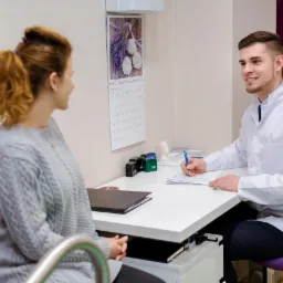 Kobieta w szarym swetrze konsultuje się z mężczyzną w białym fartuchu lekarskim, który wypełnia dokumenty przy biurku w gabinecie z kalendarzem z motywem lawendy na ścianie.