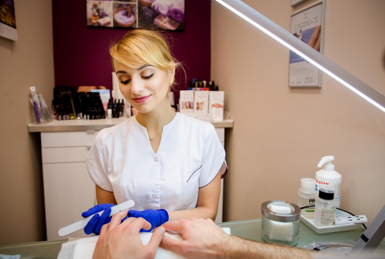 Manikiurzystka w niebieskich rękawiczkach opiłowuje paznokcie klientowi w salonie kosmetycznym, na stole leżą akcesoria do manicure, a w tle widać certyfikat i kosmetyki.