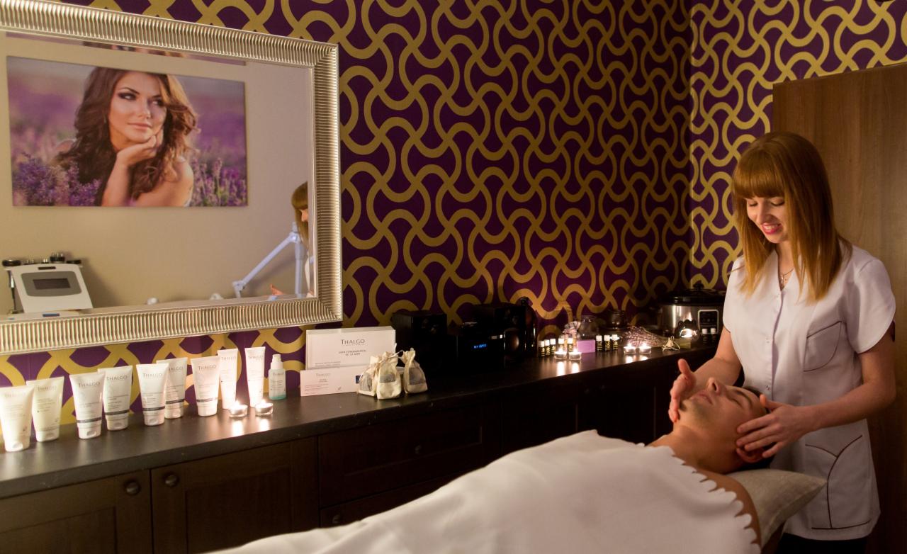 Mężczyzna relaksuje się podczas masażu twarzy w gabinecie z fioletowo-złotą tapetą, gdzie na półce widoczne kosmetyki marki Thalgo i świece.