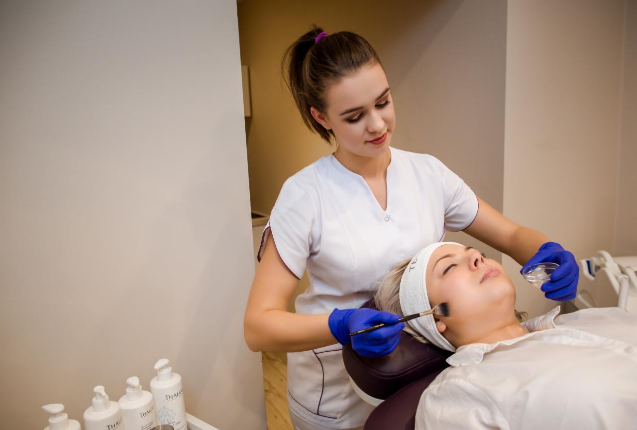 Kosmetolog w niebieskich rękawiczkach nakłada pędzlem preparat na twarz klientki leżącej na fotelu, w tle widoczne butelki kosmetyków.
