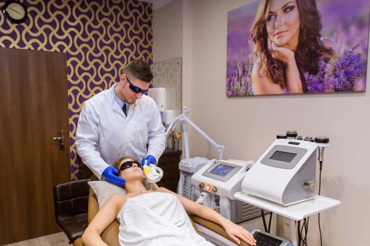 Osoba w białym fartuchu i ochronnych okularach wykonuje zabieg na twarzy klientki leżącej na fotelu, przy użyciu urządzenia kosmetycznego. W tle widoczny wzorzysty panel i obraz z motywem lawendy.