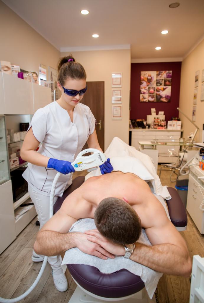 Klient leży na stole zabiegowym podczas depilacji laserowej pleców, wykonywanej przez kosmetyczkę w okularach ochronnych i niebieskich rękawiczkach. W tle widoczne wyposażenie gabinetu.