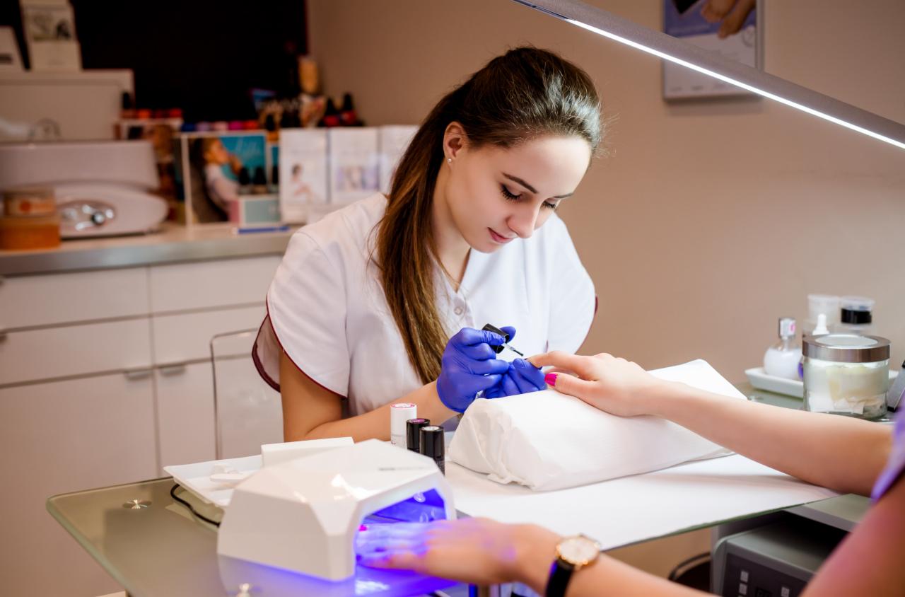 Kobieta w niebieskich rękawiczkach maluje paznokcie klientki na różowo w dobrze oświetlonym gabinecie, widoczna lampa UV do utwardzania lakieru.