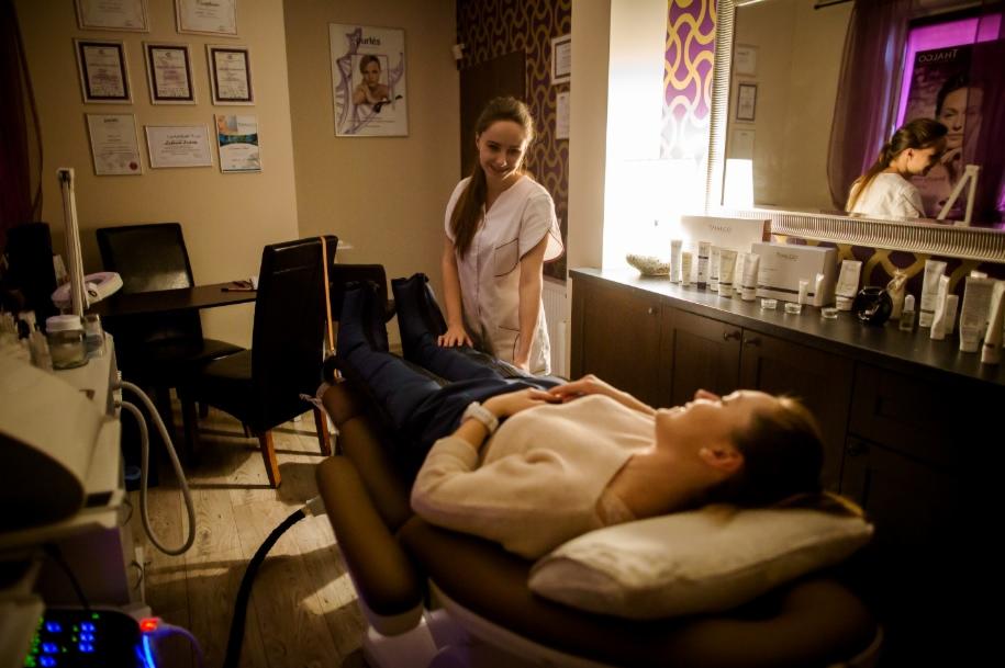 Klientka leży na stole do zabiegów z nogami w specjalnych mankietach, uśmiechnięta pracownica w białym uniformie obok, w tle ściana z dyplomami i półka z kosmetykami.